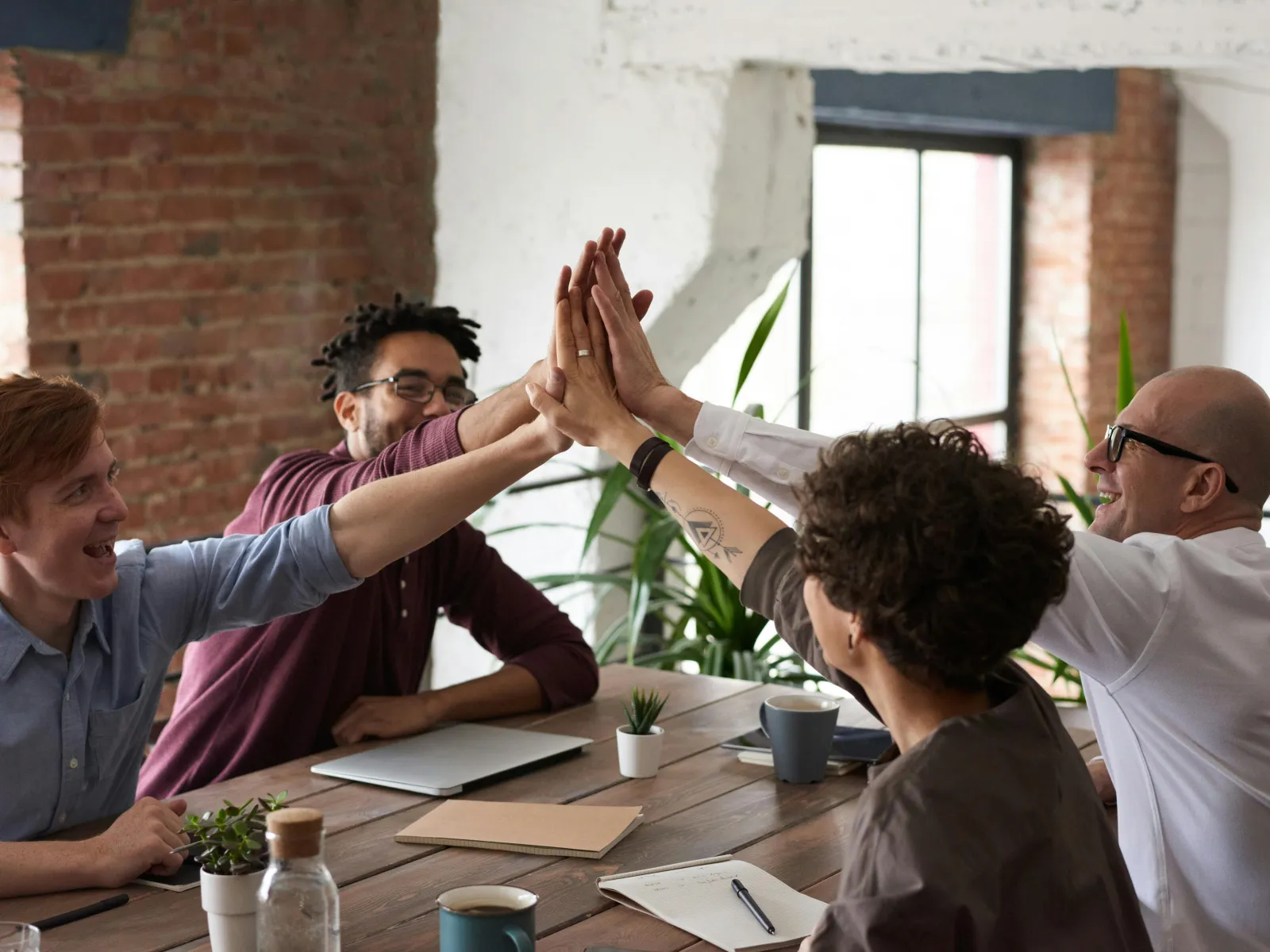 Four diverse coworkers in a modern office high-fiving over a wooden table with laptops and plants.