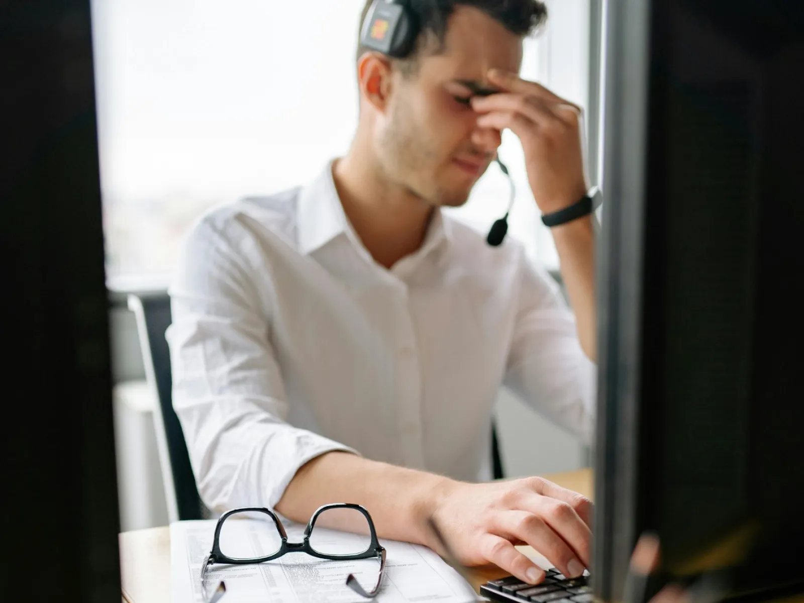 Stressed man wearing headset at office desk with computer, glasses on documents, and pen holder in foreground