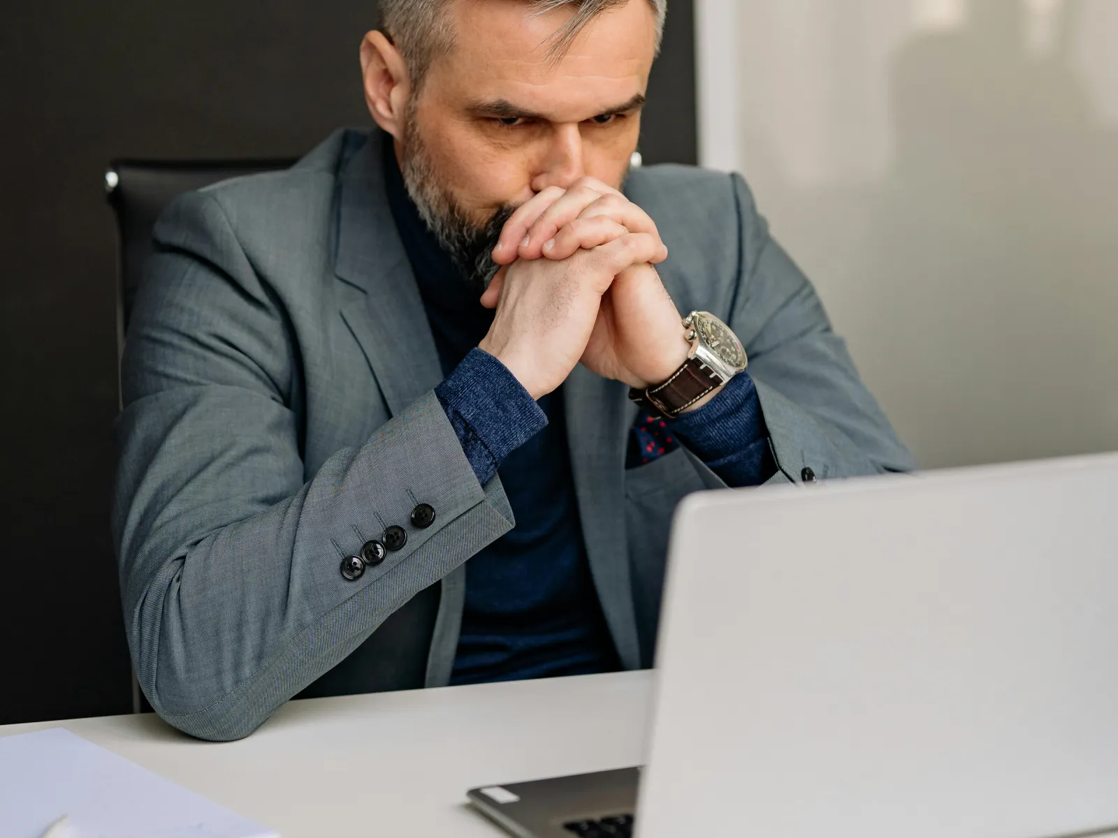 Thoughtful businessman in gray suit staring at laptop with hands clasped in front of his face in modern office.