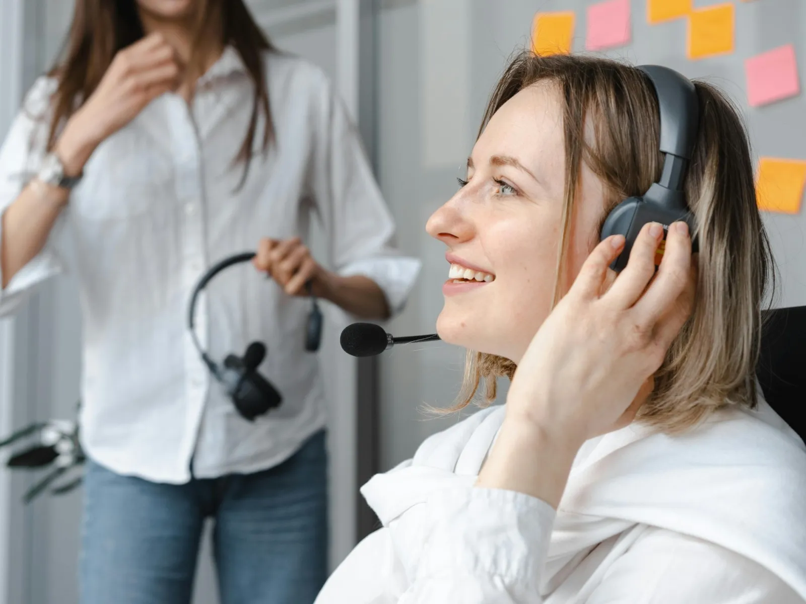 Smiling woman wearing headset with microphone in office setting with colleague and sticky notes in background