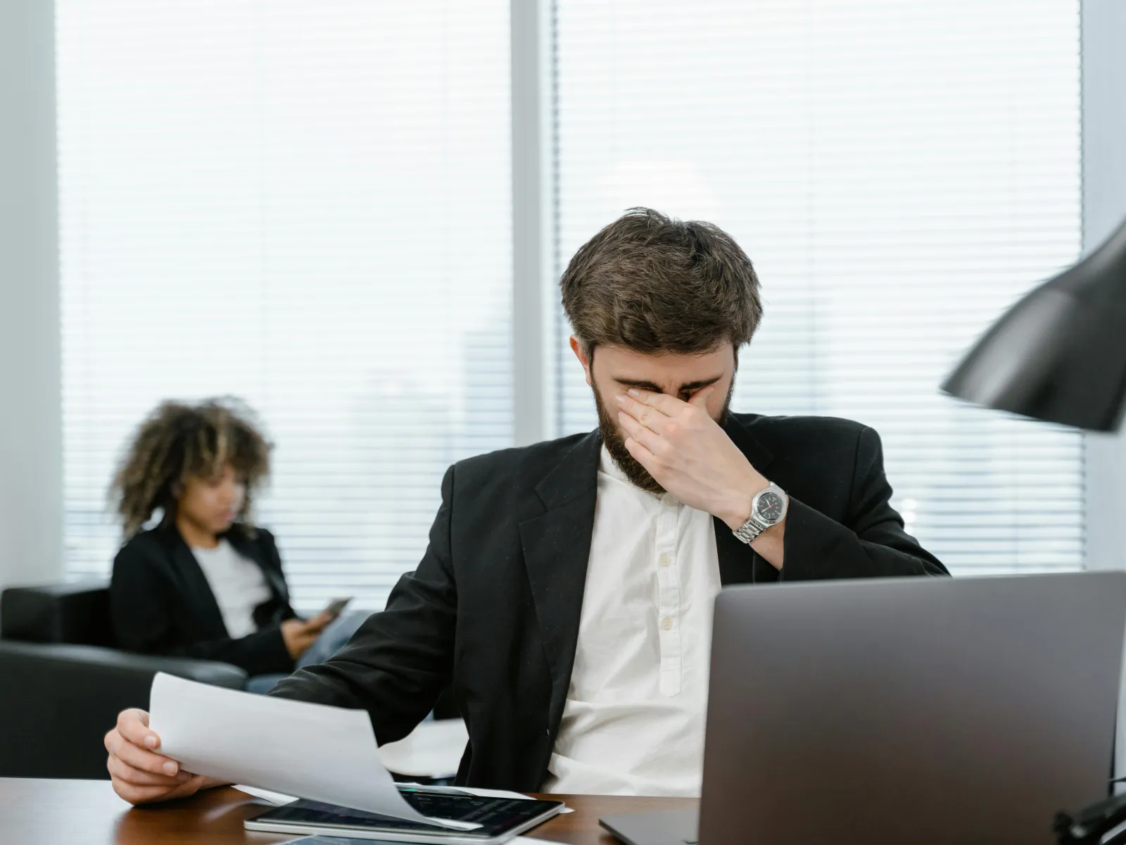 Stressed businessman in suit holding paperwork and pinching his nose bridge at office desk with laptop