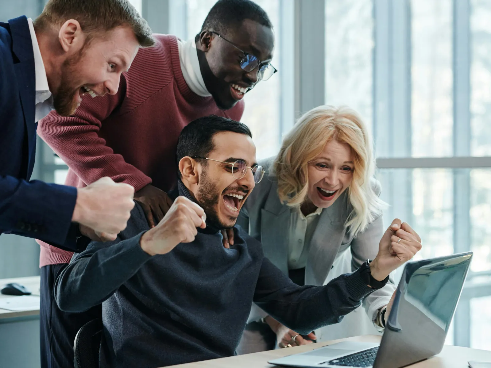 Four diverse coworkers excitedly celebrate success while looking at a laptop in a bright modern office.