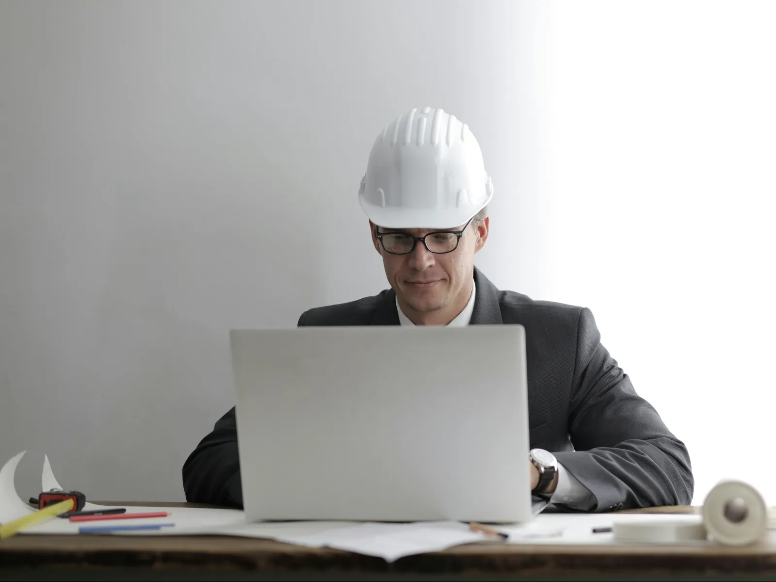 Engineer in white hard hat and glasses working on laptop at desk with blueprints and tools.
