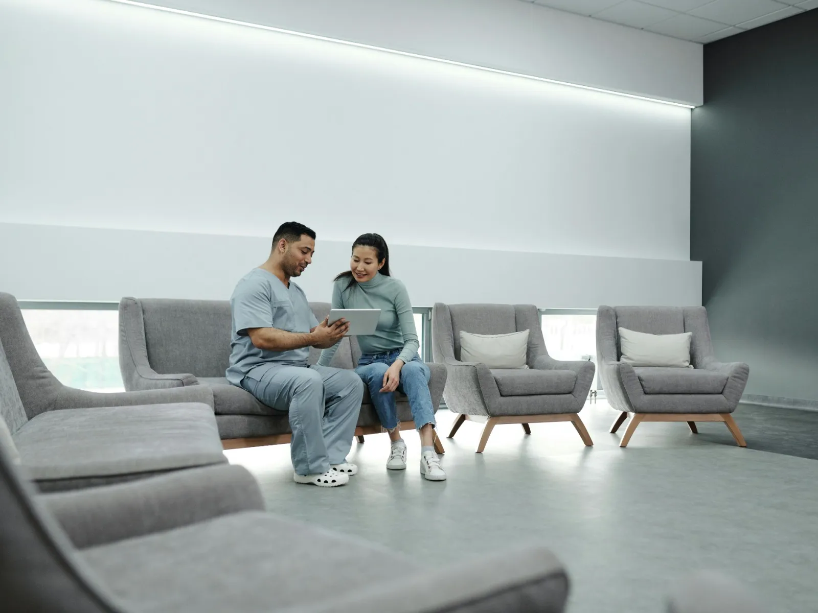 Medical professional and patient reviewing information on a tablet in a modern waiting area with gray chairs