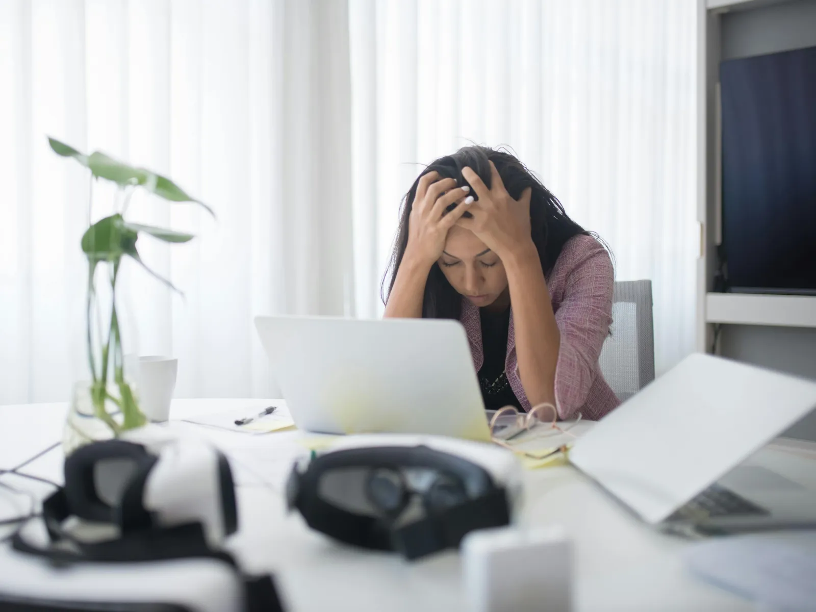 Stressed woman holding her head working on laptop in a bright office with headphones and plants nearby.