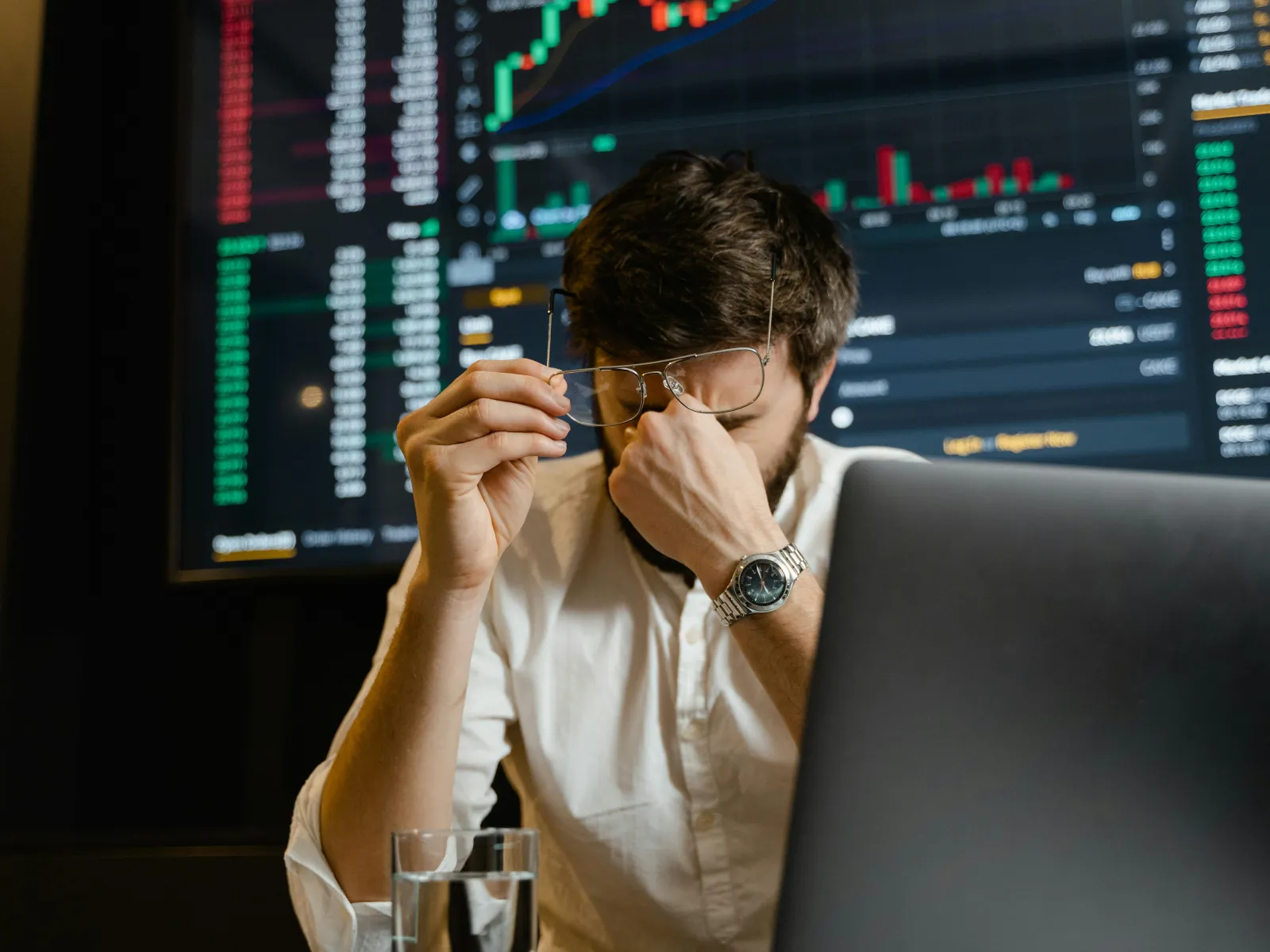 Stressed man in white shirt holding glasses, rubbing eyes in front of laptop with financial stock charts in background
