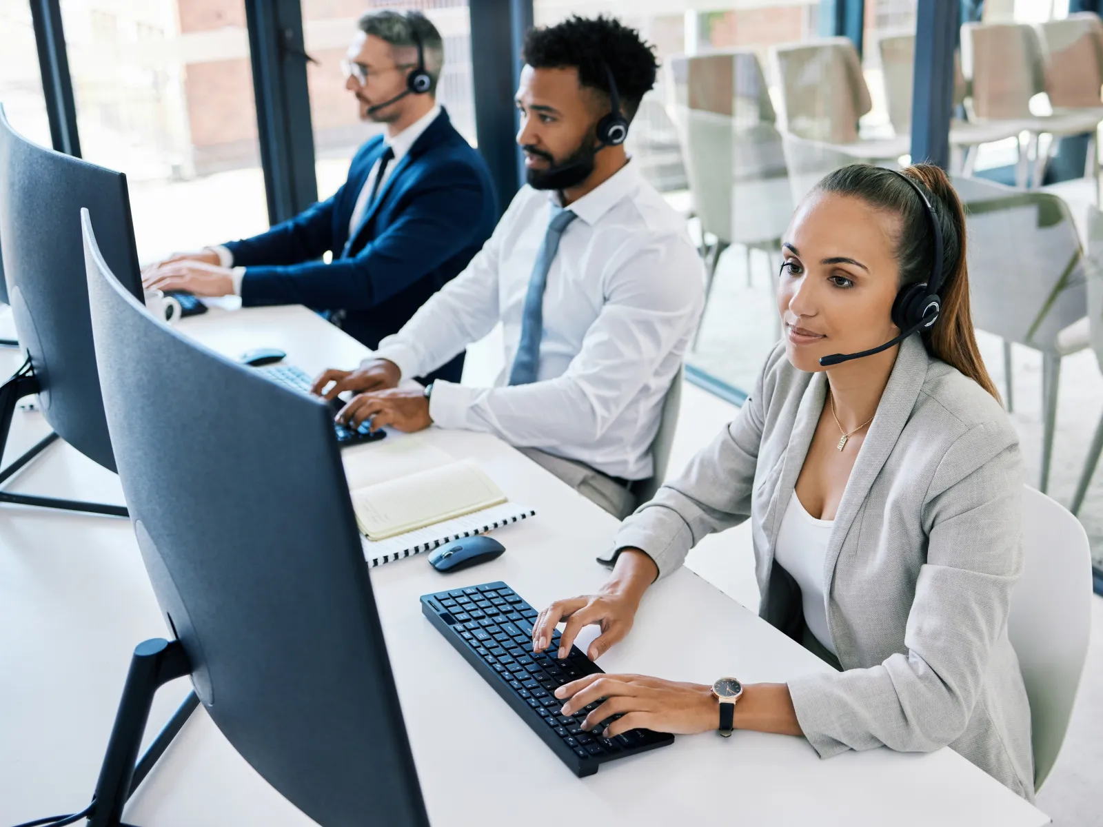 Call center team wearing headsets working on computers in a modern office environment with large windows.