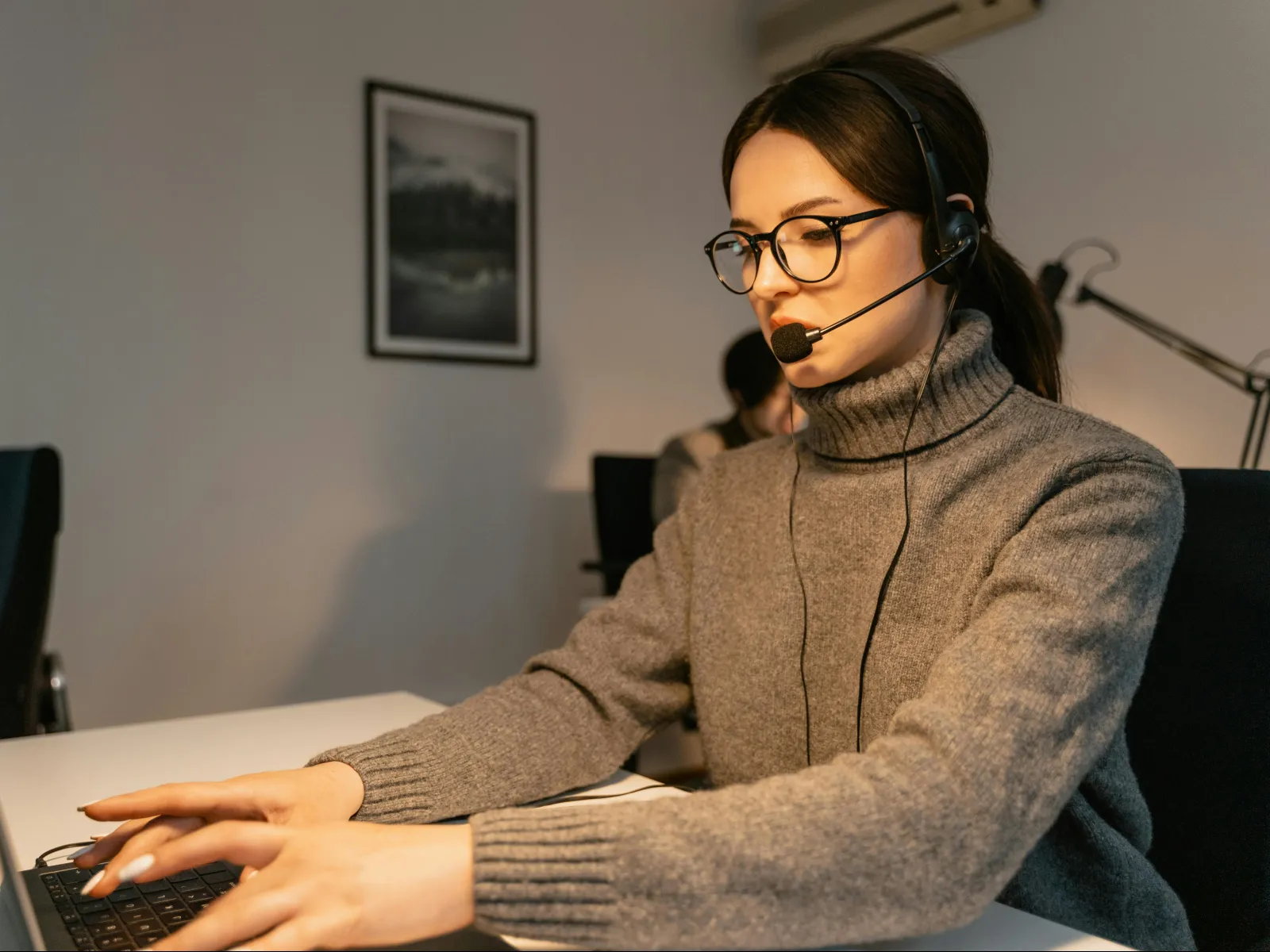 Woman wearing headset and glasses working on laptop in modern office setting with dim lighting.