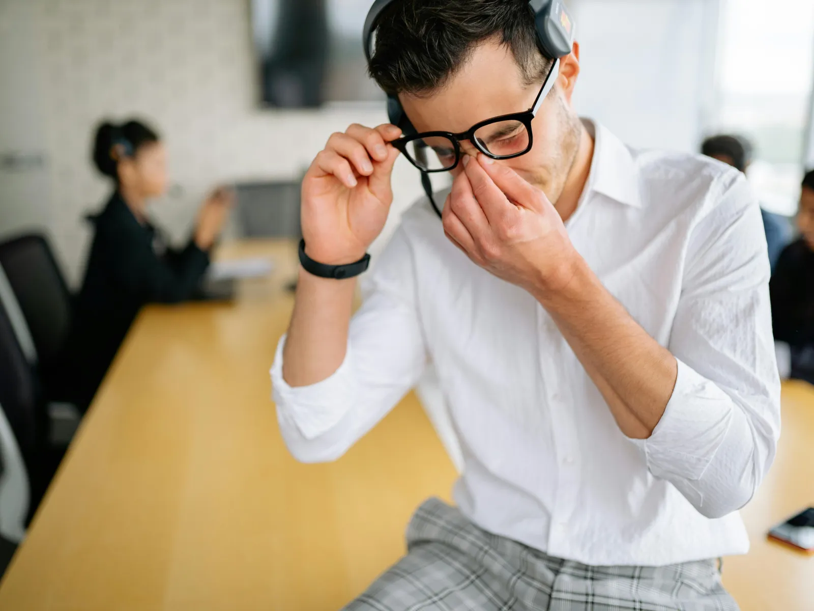Stressed man in glasses and headset rubbing his eyes in office meeting room with colleagues in background.