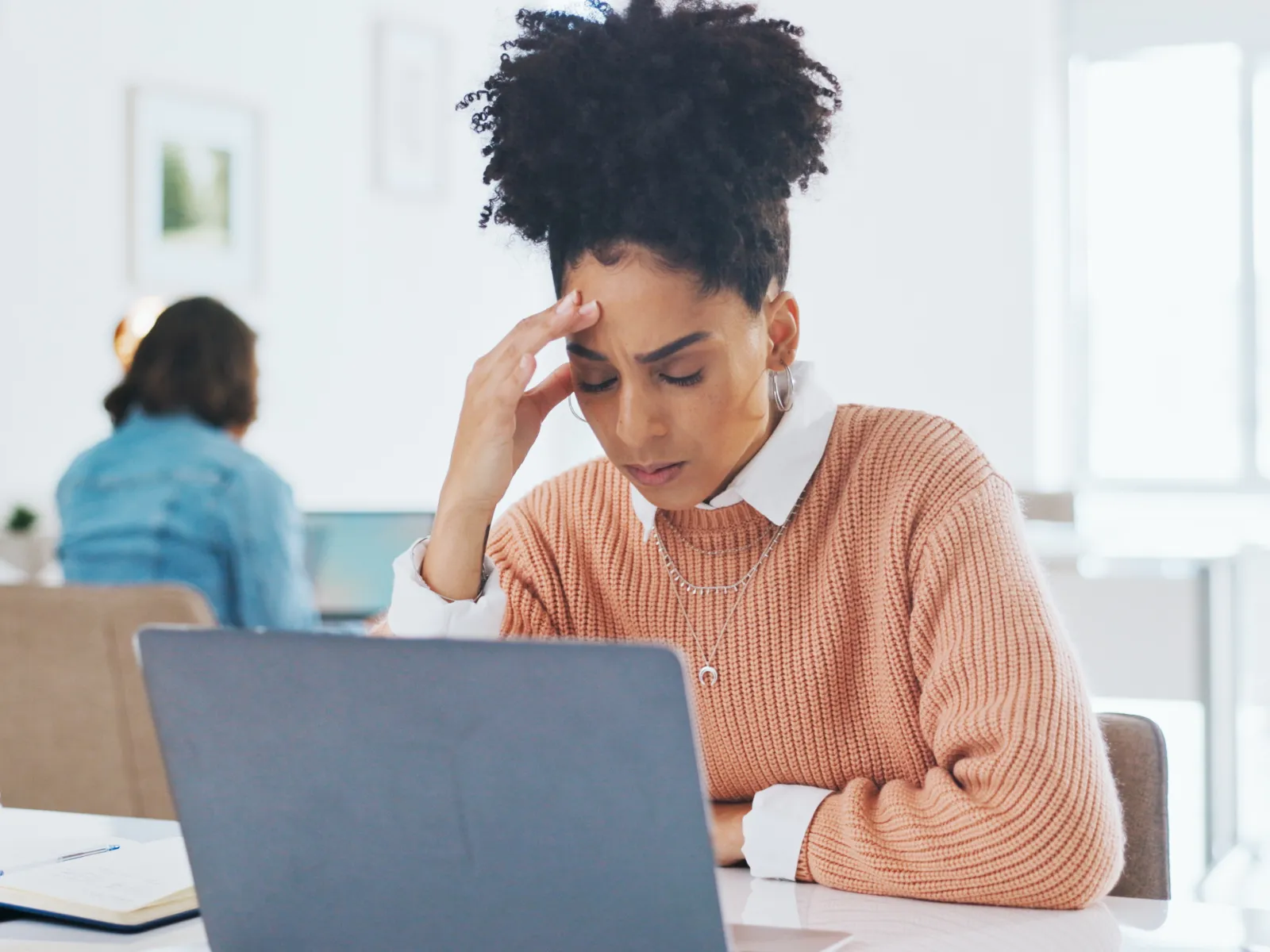 Woman in pink sweater working on laptop looking stressed in bright office with coffee cup and notebook nearby