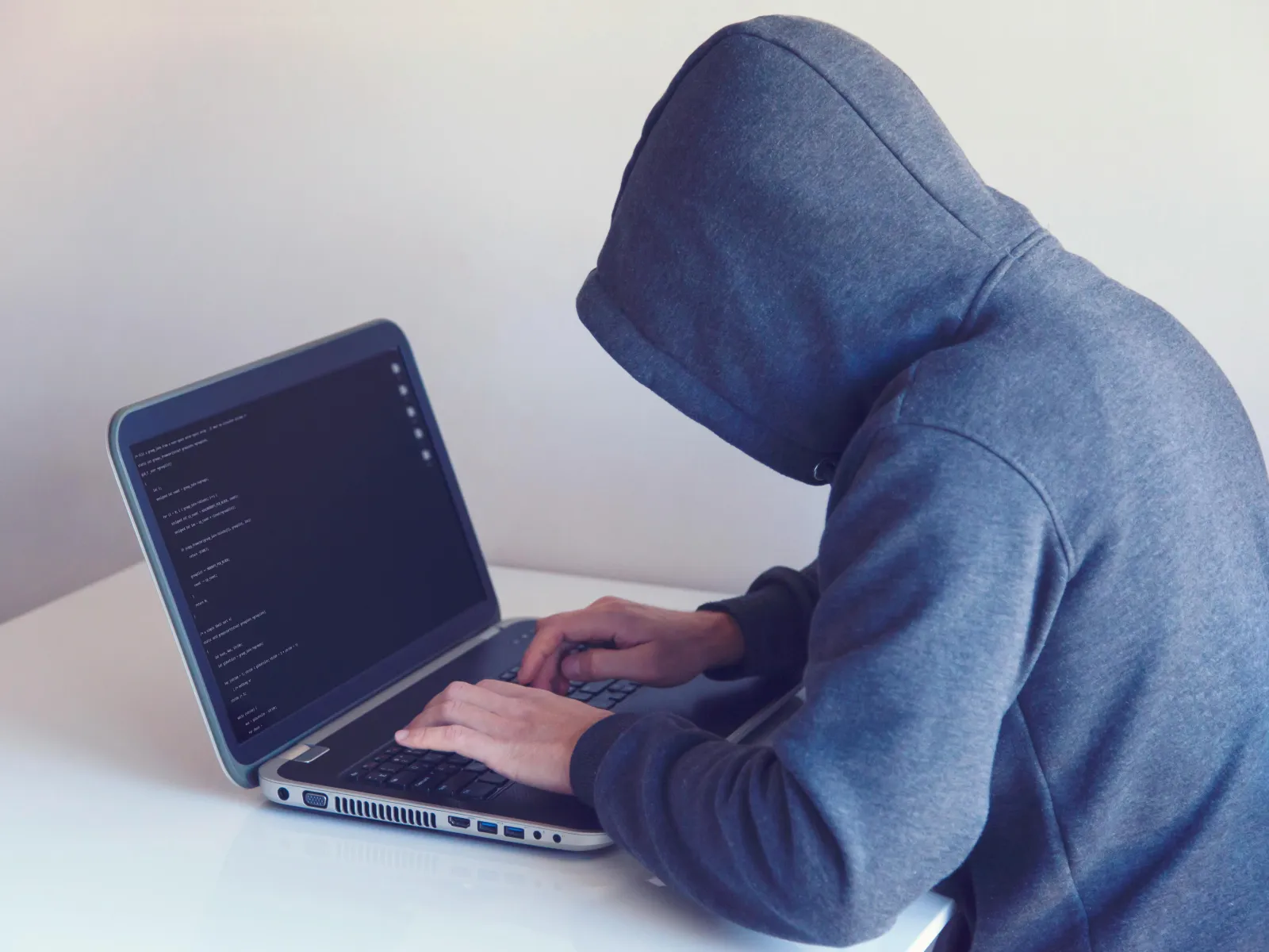 Person in gray hoodie typing on a laptop with dark screen displaying code, seated at a white desk.