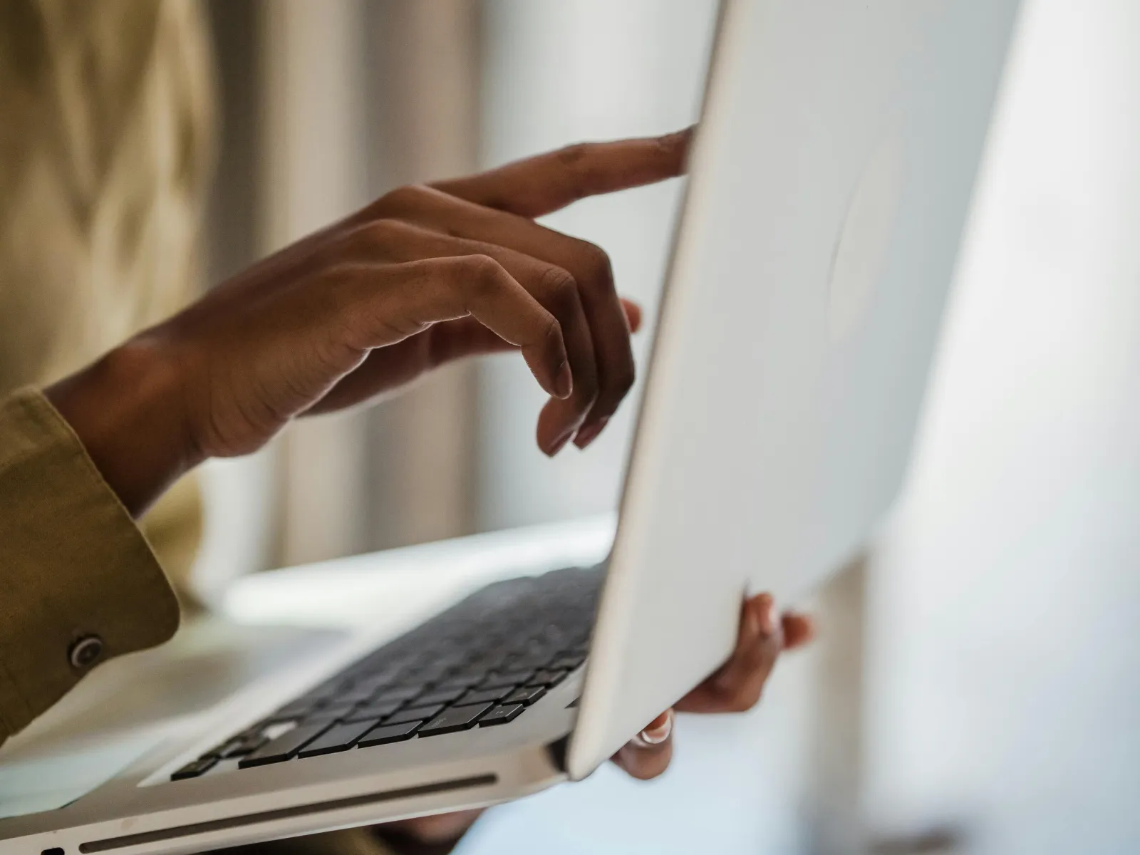 Person holding a laptop with one hand and pointing at the screen with the other in a bright room.