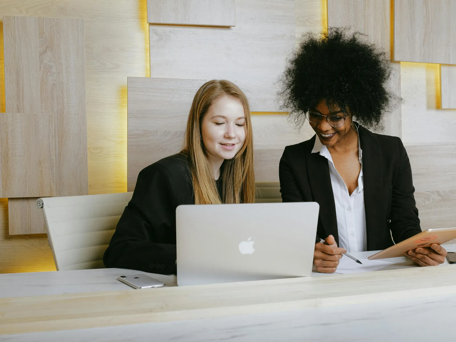 Two professional women collaborating over a laptop and documents in a modern office setting.