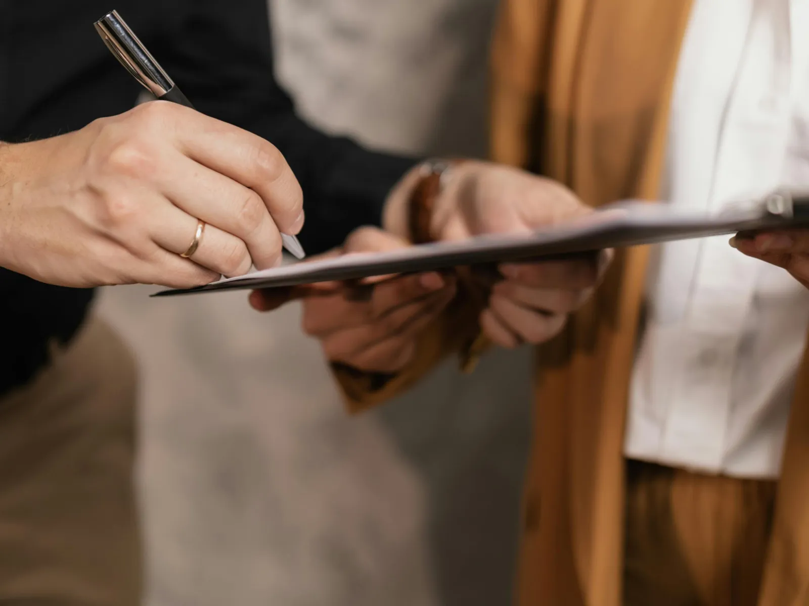 Man signing document on clipboard while woman in brown suit holds it, close-up of hands and paperwork