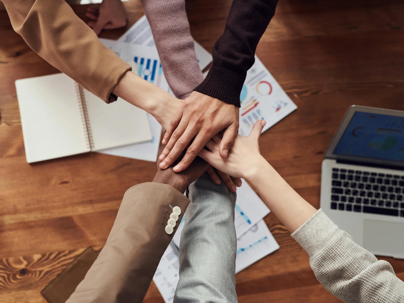Diverse team joining hands over a wooden table with laptop and business documents symbolizing teamwork and collaboration.