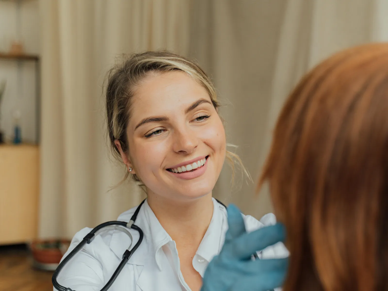 Smiling female doctor wearing gloves and a stethoscope examining a patient in a medical office.