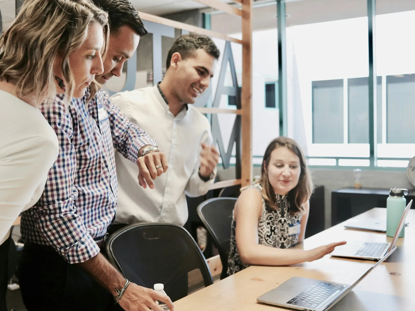 Four professionals collaborating and discussing work at a modern office desk with laptops and a water bottle.