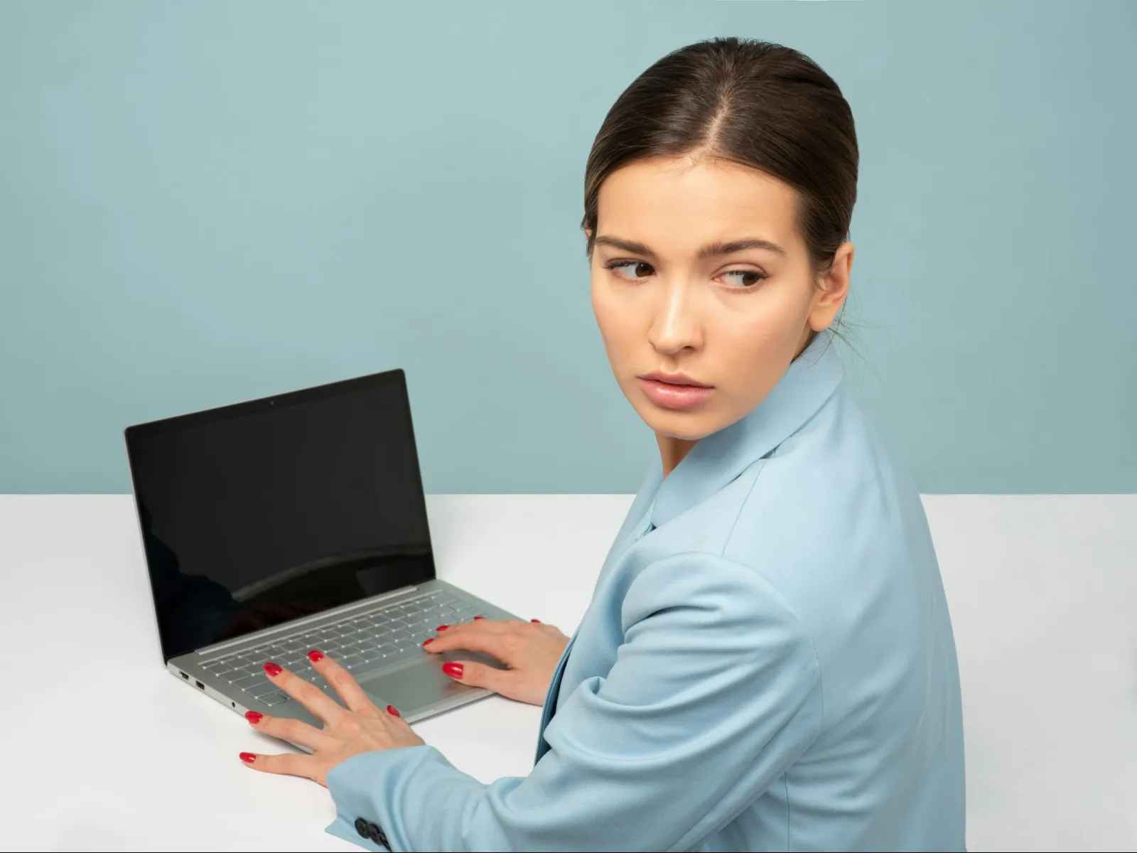 Young woman in blue blazer looking over shoulder while using a laptop on a white desk with pale blue background
