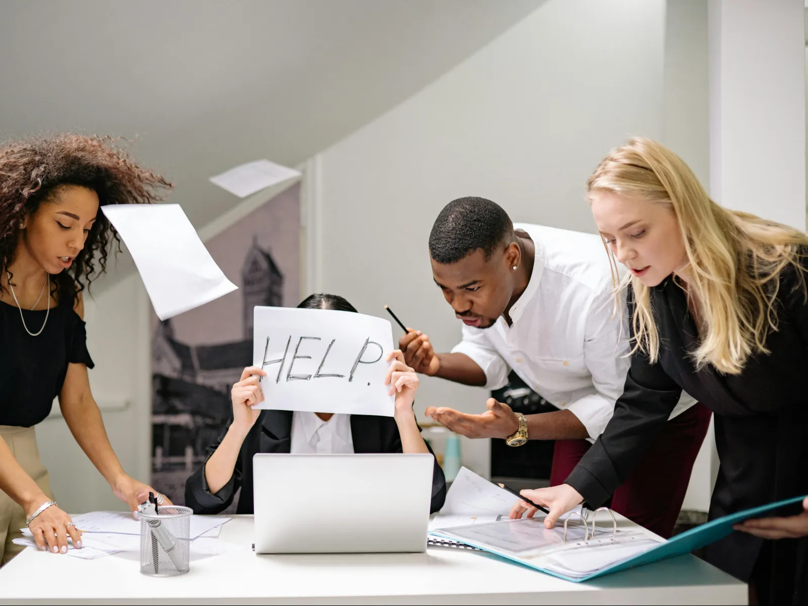 Stressed office worker holding a HELP sign with colleagues discussing documents in a busy workspace.