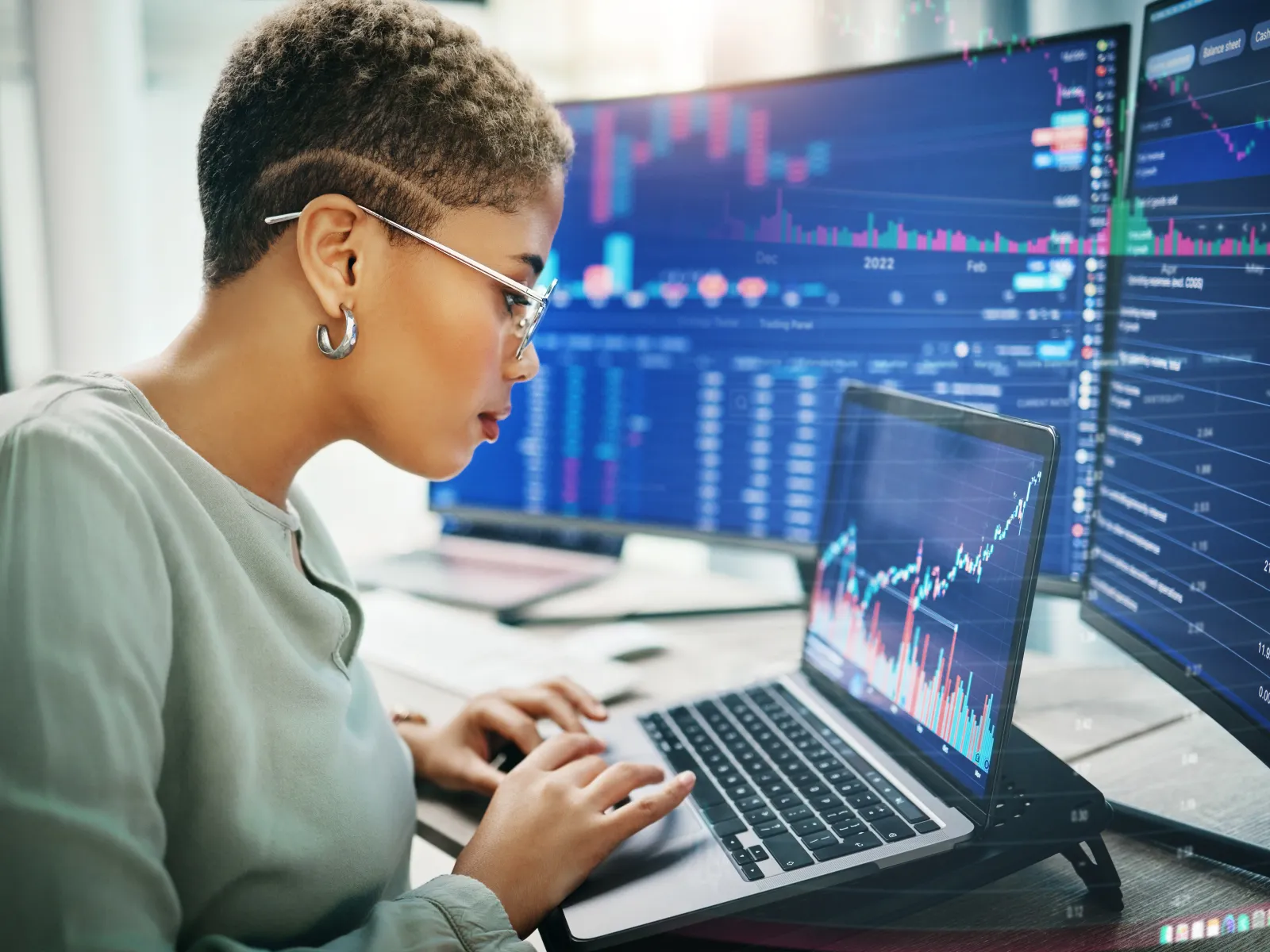 Woman analyzing stock market data on laptop with multiple large monitors displaying financial charts and graphs.