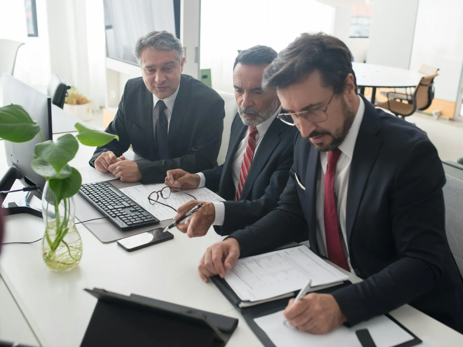Three businessmen in suits collaborating over documents and a tablet in a modern office setting.
