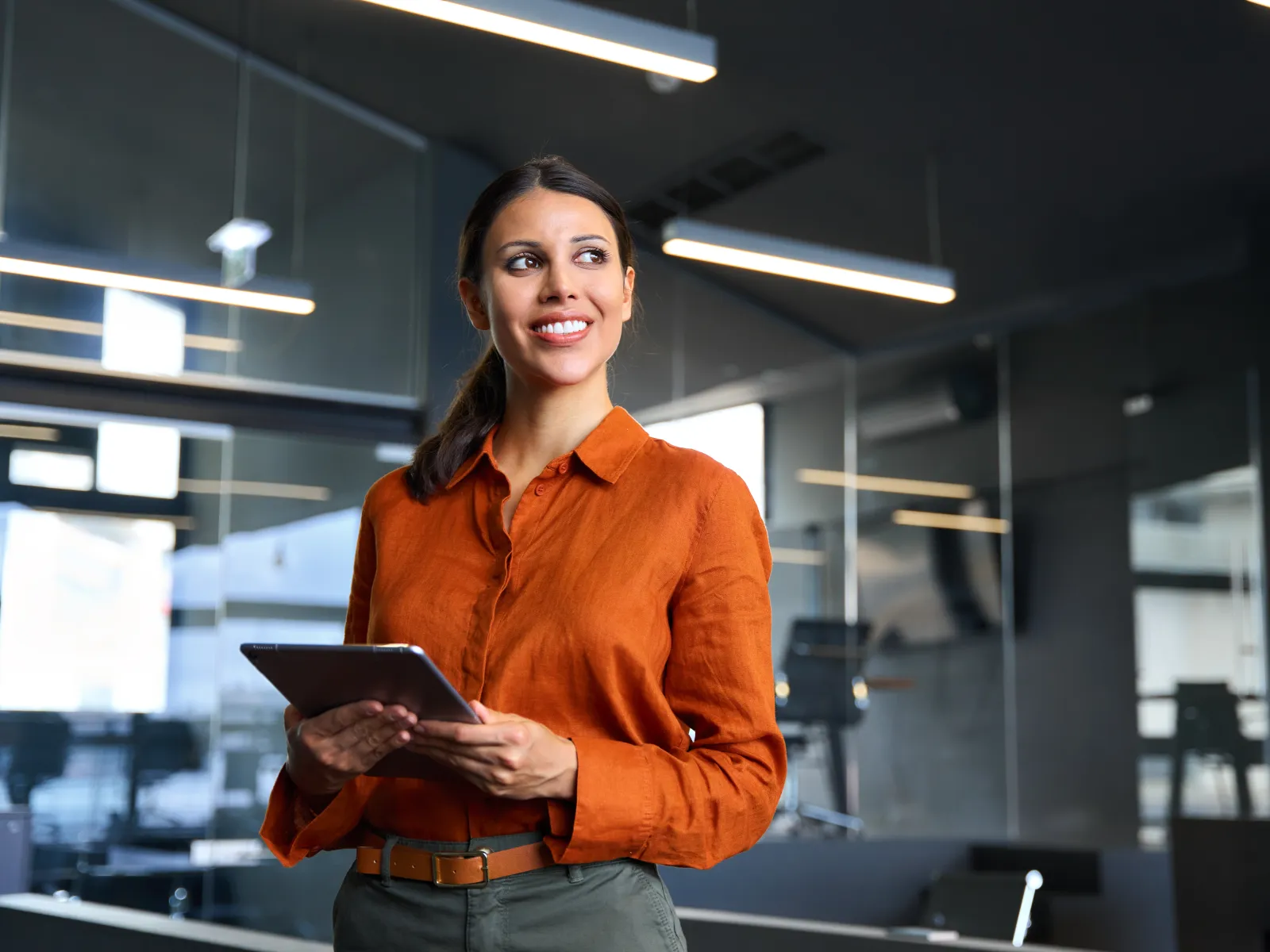 Confident woman in orange shirt holding tablet in modern office with glass walls and ceiling lights