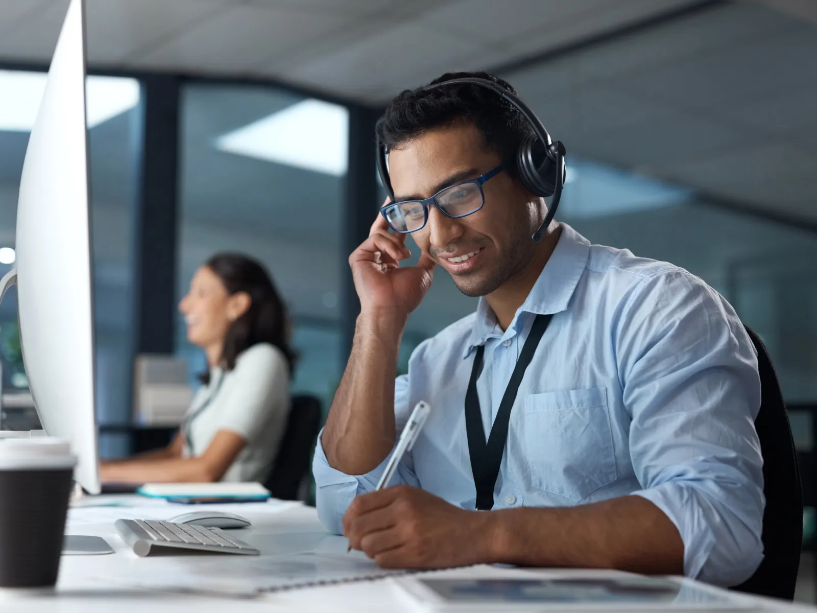 Man wearing headset and glasses working at a computer in modern office with a female colleague in the background