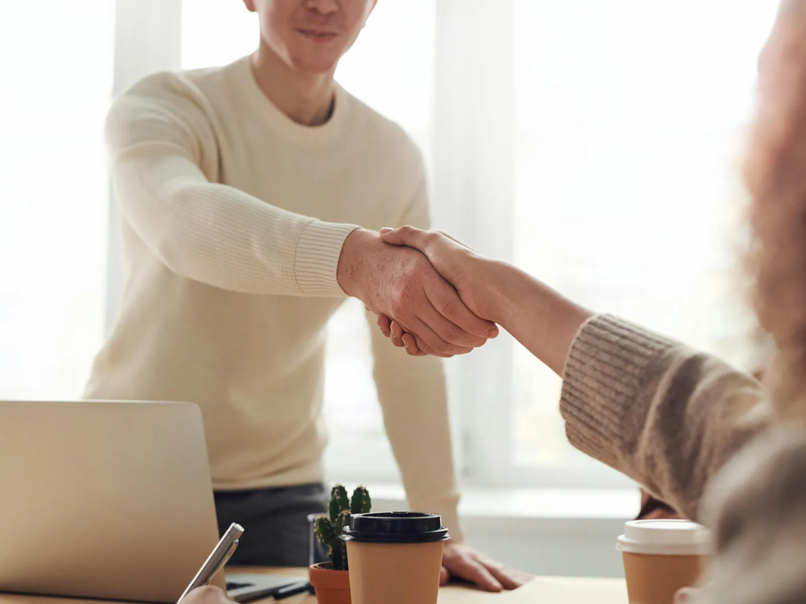 Two people shaking hands over a desk with laptops and coffee cups in a bright office setting