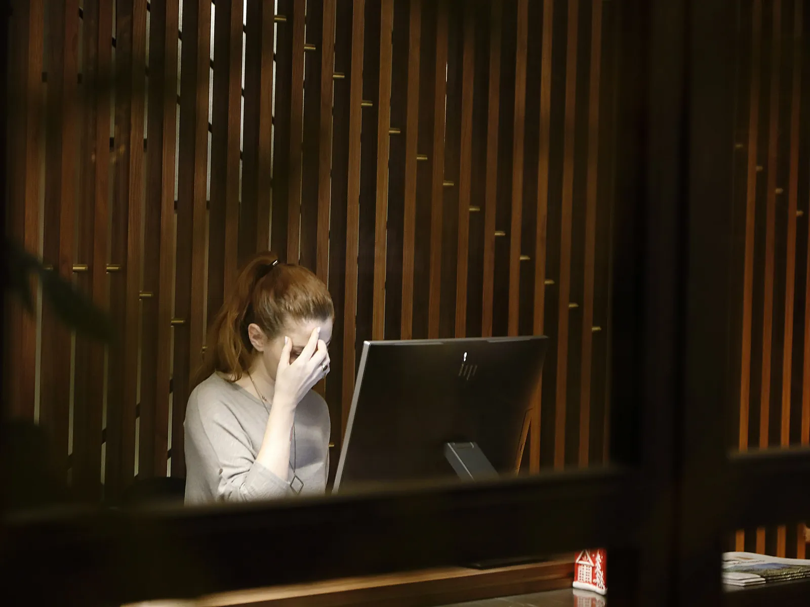 Stressed woman sitting at desk with computer in a dimly lit office with wooden panel walls.