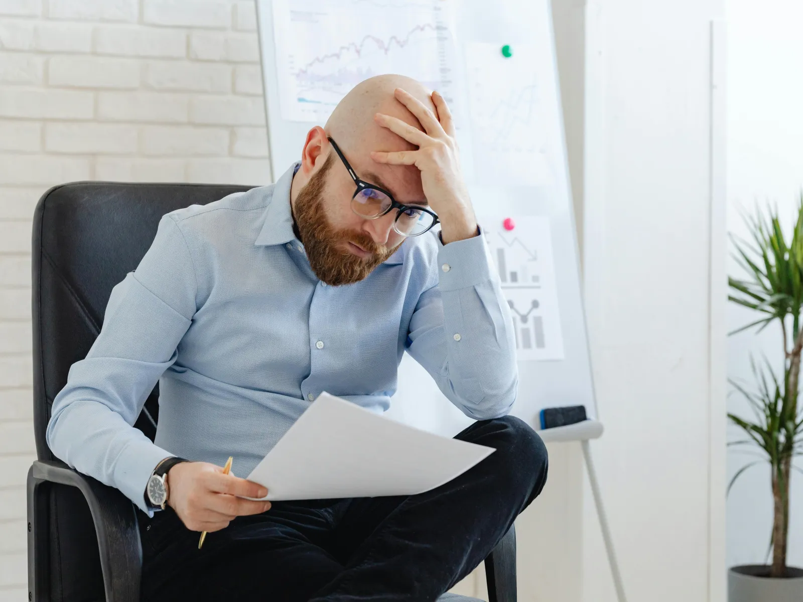 Stressed businessman in glasses reviewing documents with charts in a modern office setting