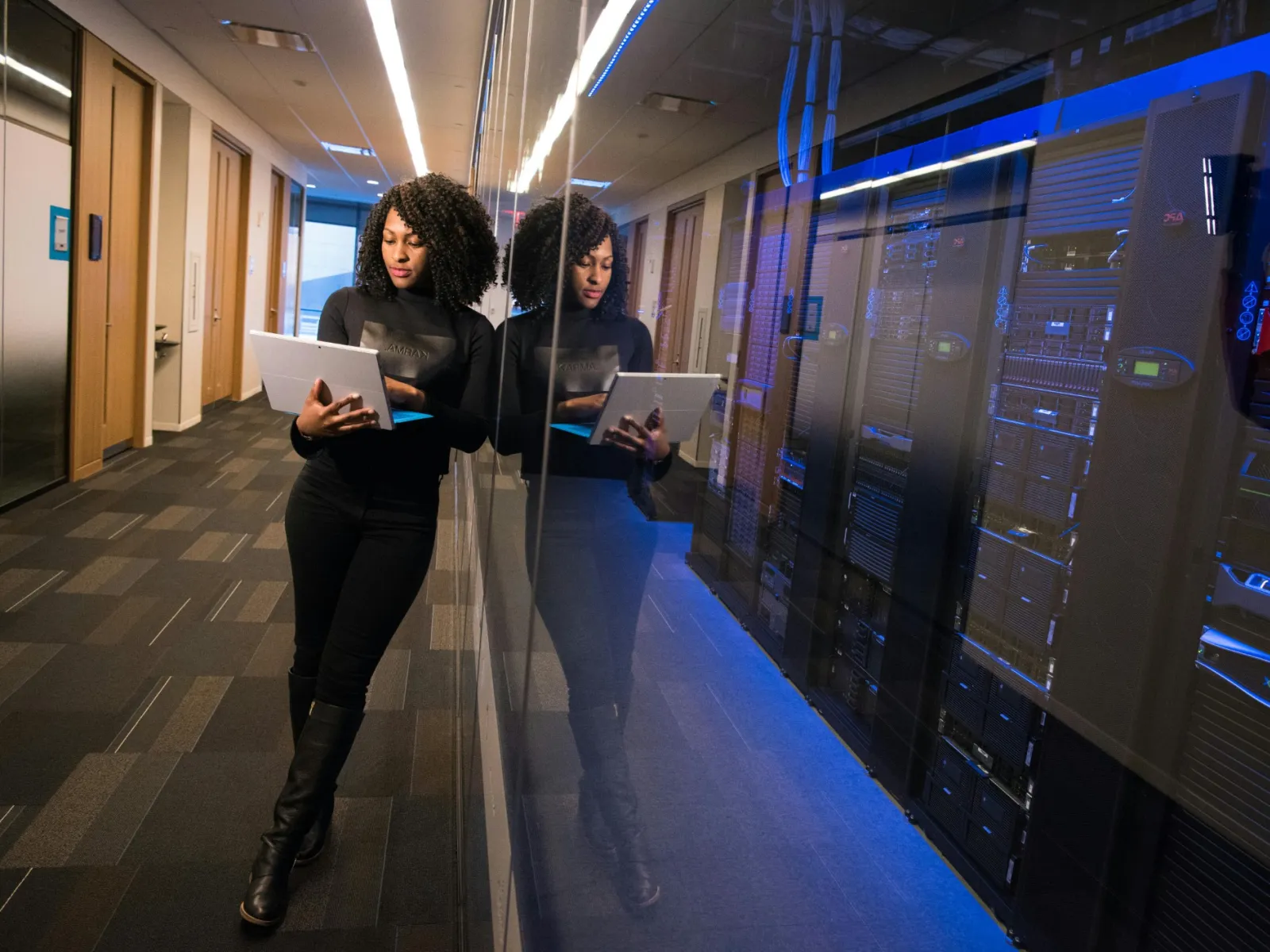 Woman in black outfit working on laptop beside server racks in a modern data center corridor.