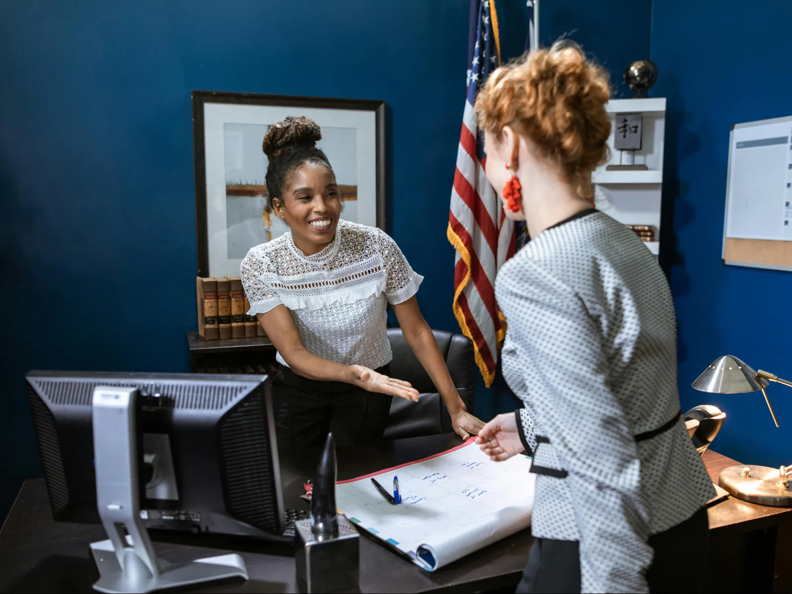 Two women discussing plans on a desk in an office with American flag and calendar in the background