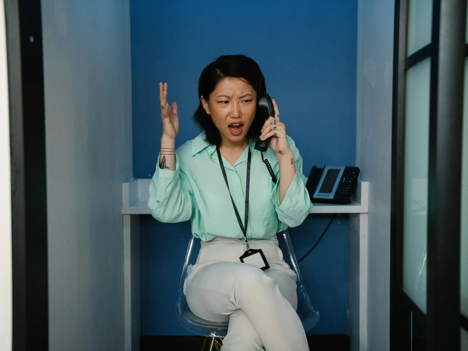 Frustrated woman in office attire talking on phone with animated expression in a small workspace.