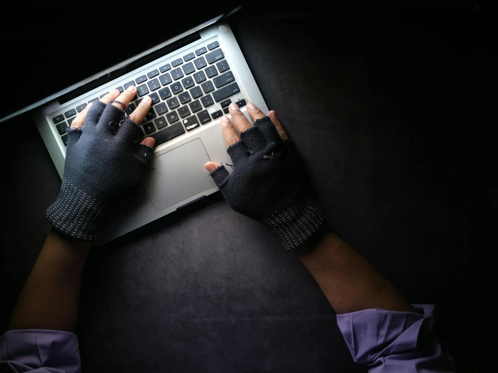 Person wearing fingerless gloves typing on a laptop keyboard on a dark surface, viewed from above.