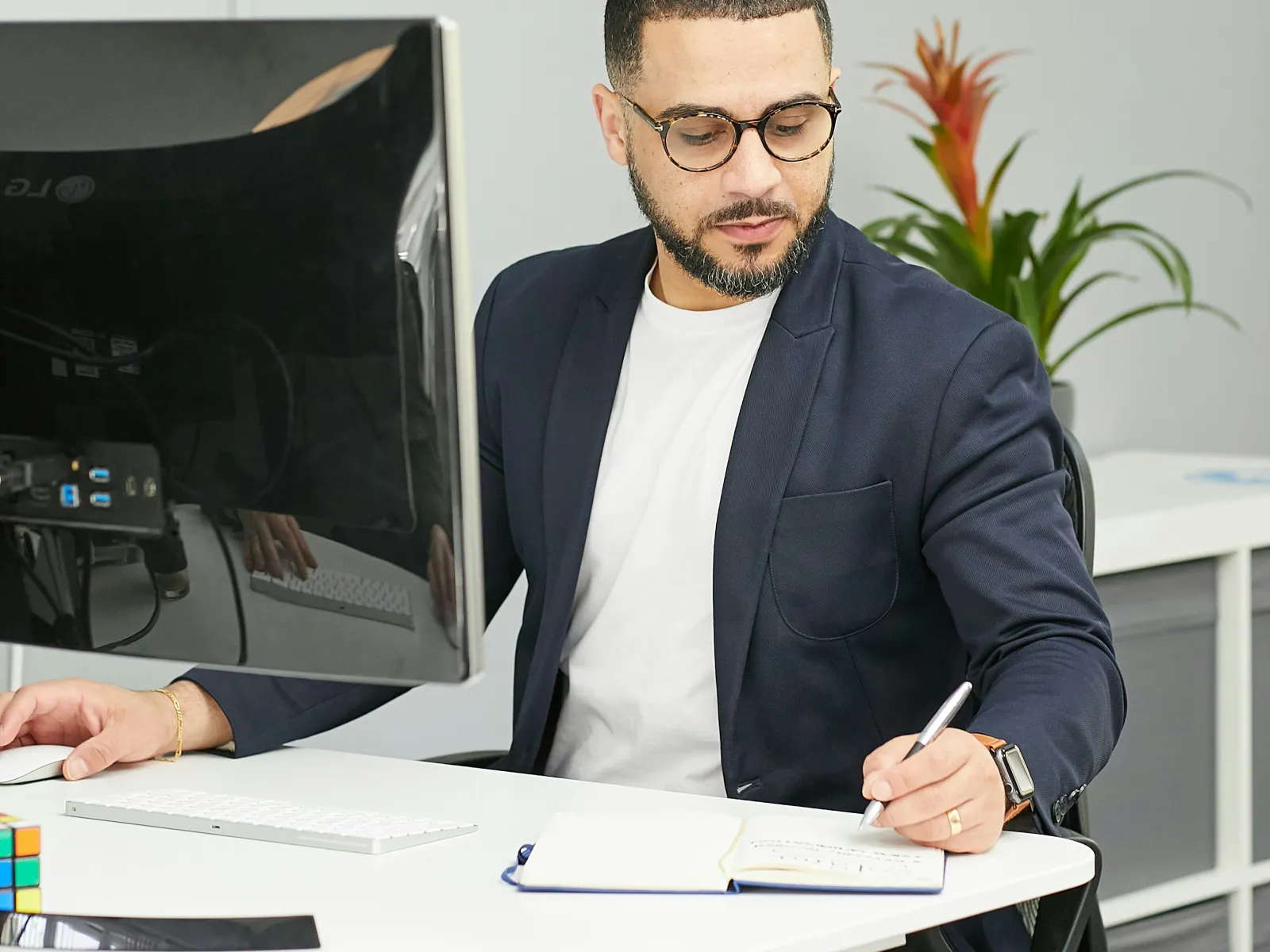 Man in glasses and navy blazer writing in a notebook at a white desk with computer and plant behind.