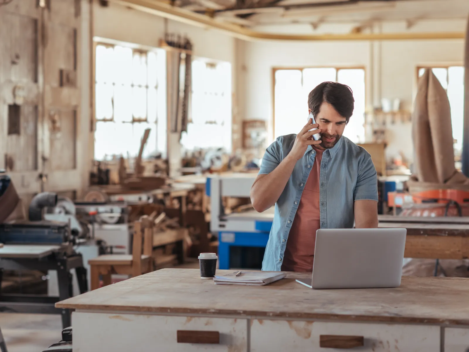 Man talking on phone while working on laptop in a bright, busy woodworking workshop with tools and machinery.