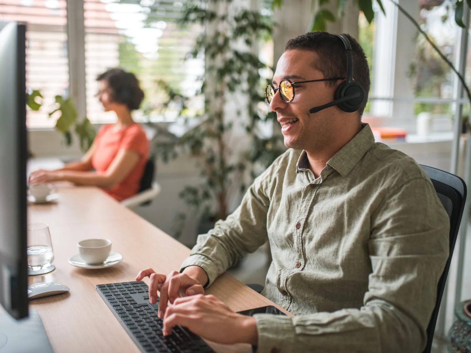 A customer service representative at a desk using a headset, with a colleague in the background.