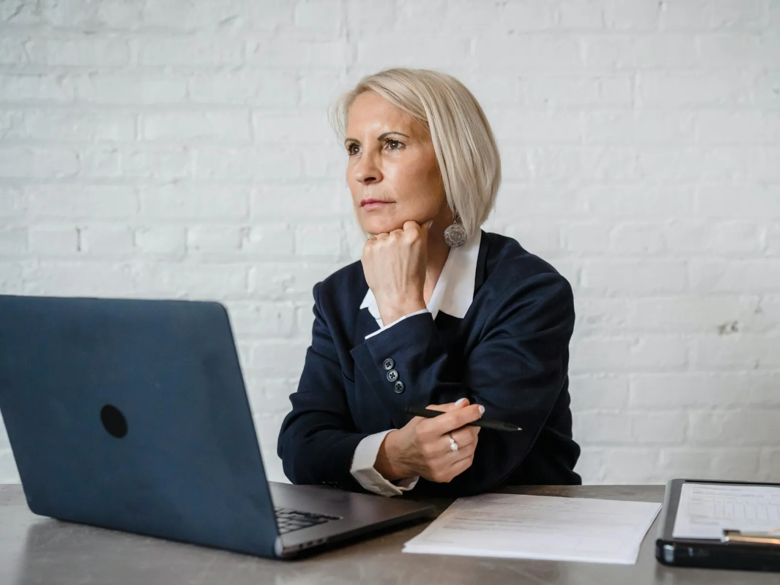 Middle-aged woman in business attire working thoughtfully on a laptop at a desk with documents and clipboard.