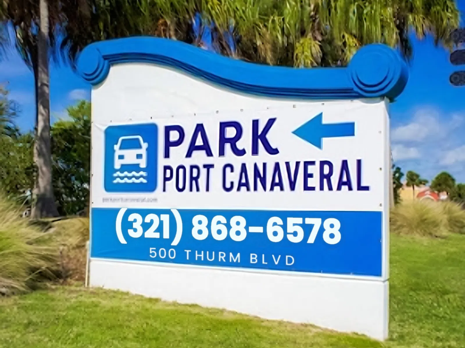 Directional sign for Park Port Canaveral with contact number and address on grassy area under blue sky.