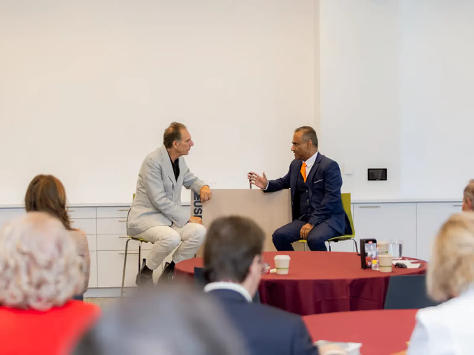 Two businessmen in suits engaged in a discussion during a professional conference with an audience watching.