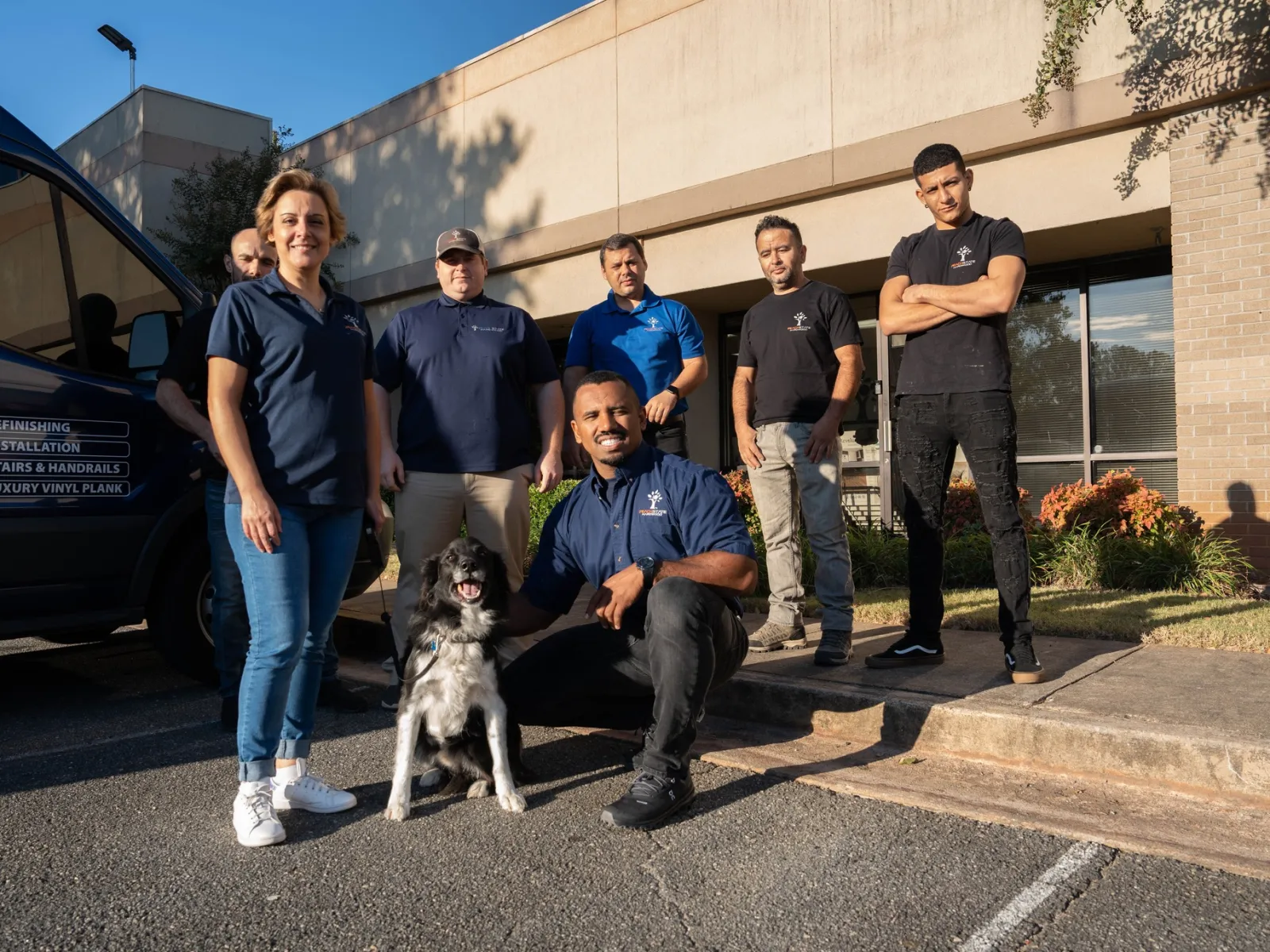 Group of six workers and a dog posing outside a building near a work van on a sunny day