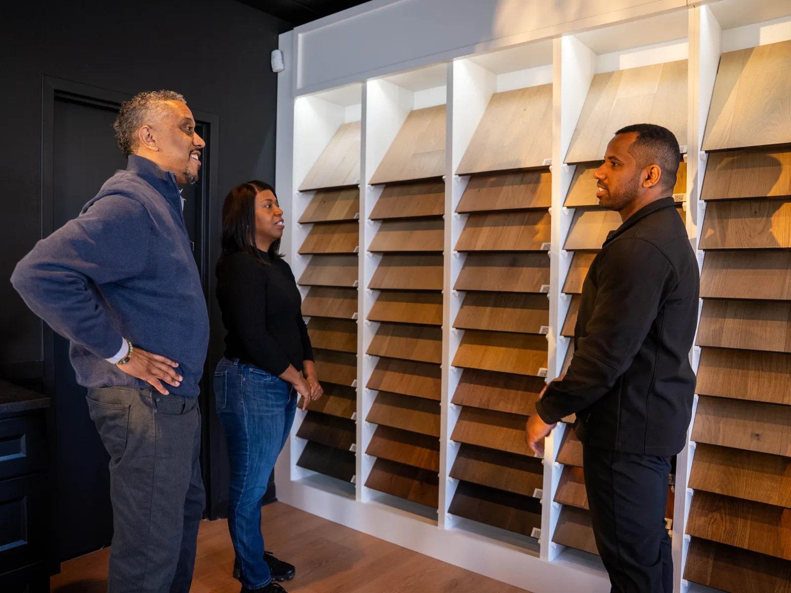 Three people discussing wood flooring samples displayed on a wall in a showroom setting.