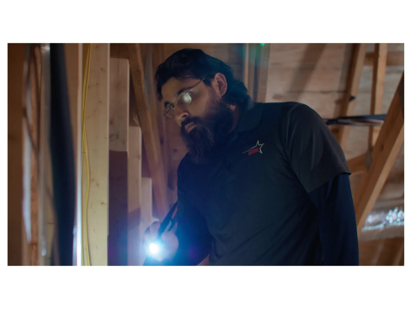 Man with glasses and beard inspecting wooden beams inside attic using flashlight in low light.