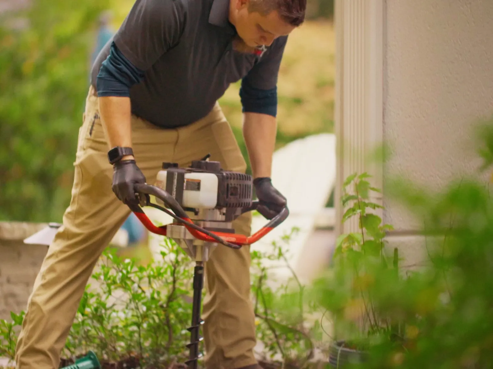 Man using a gas-powered auger to drill into soil near a house exterior during landscaping work.