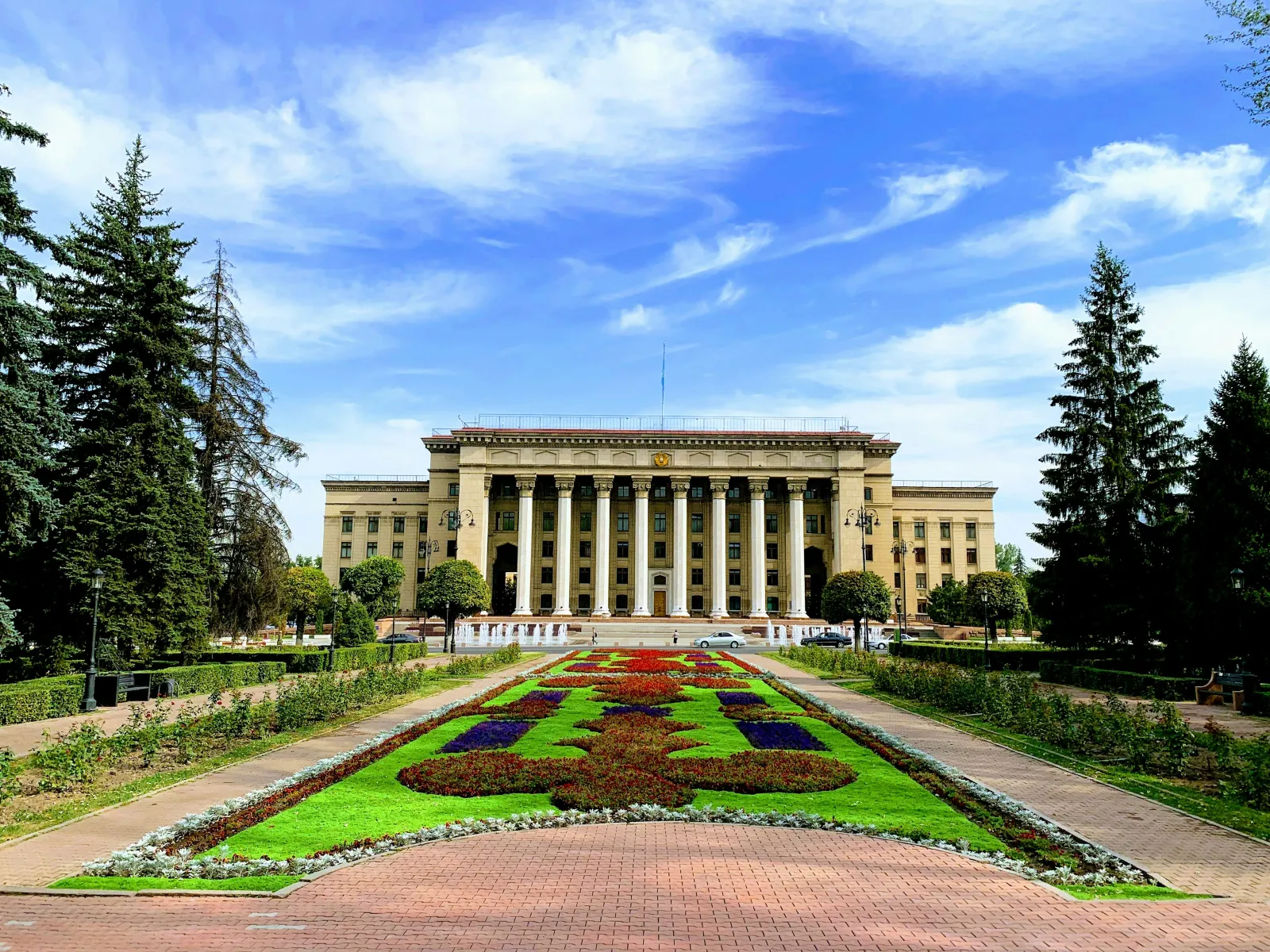 Grand neoclassical building with columns behind a colorful flower garden and pathways under a partly cloudy sky.