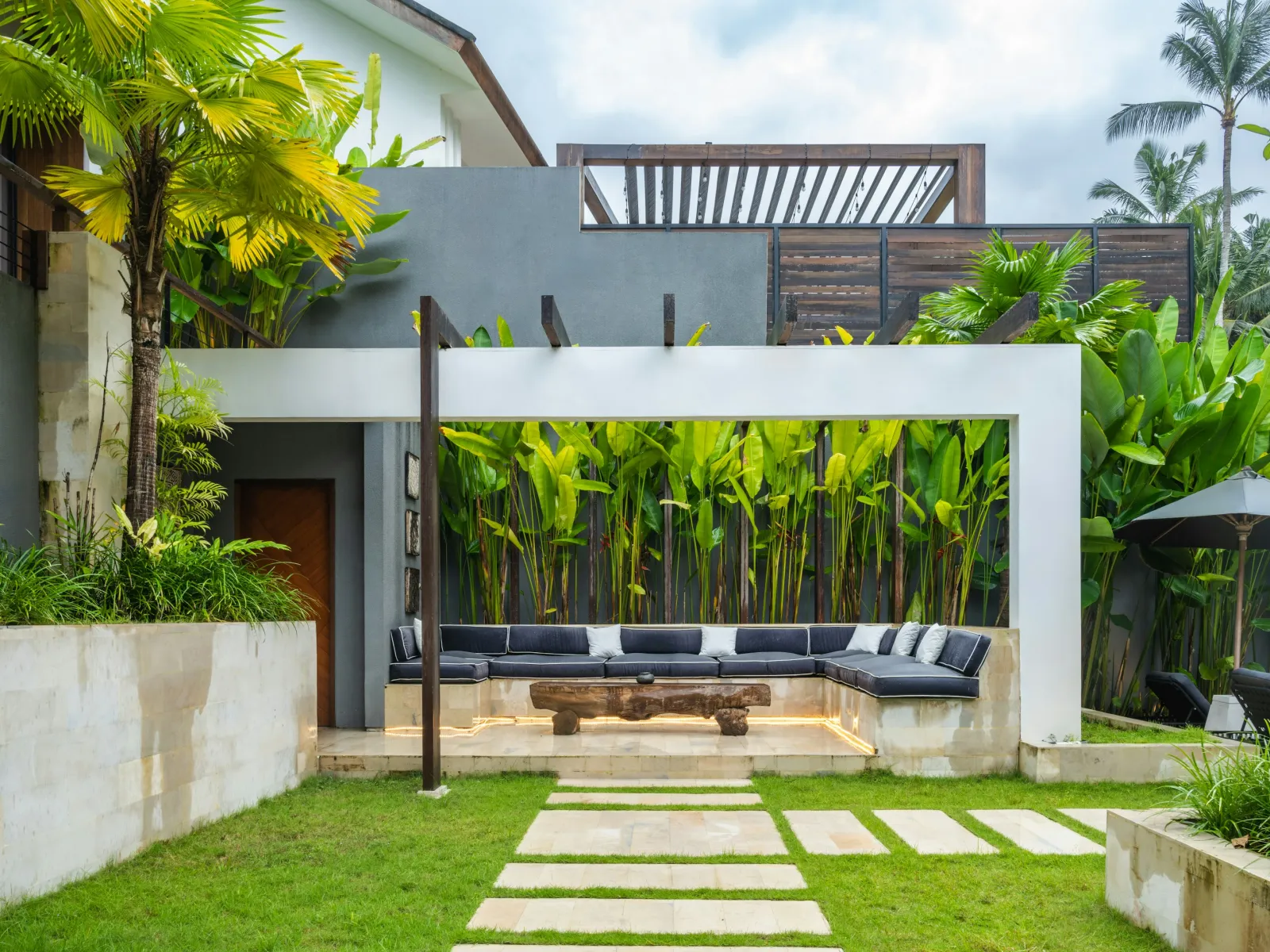 Modern outdoor patio with built-in sectional sofa, wooden table, stone pathway, and tropical greenery in backyard.