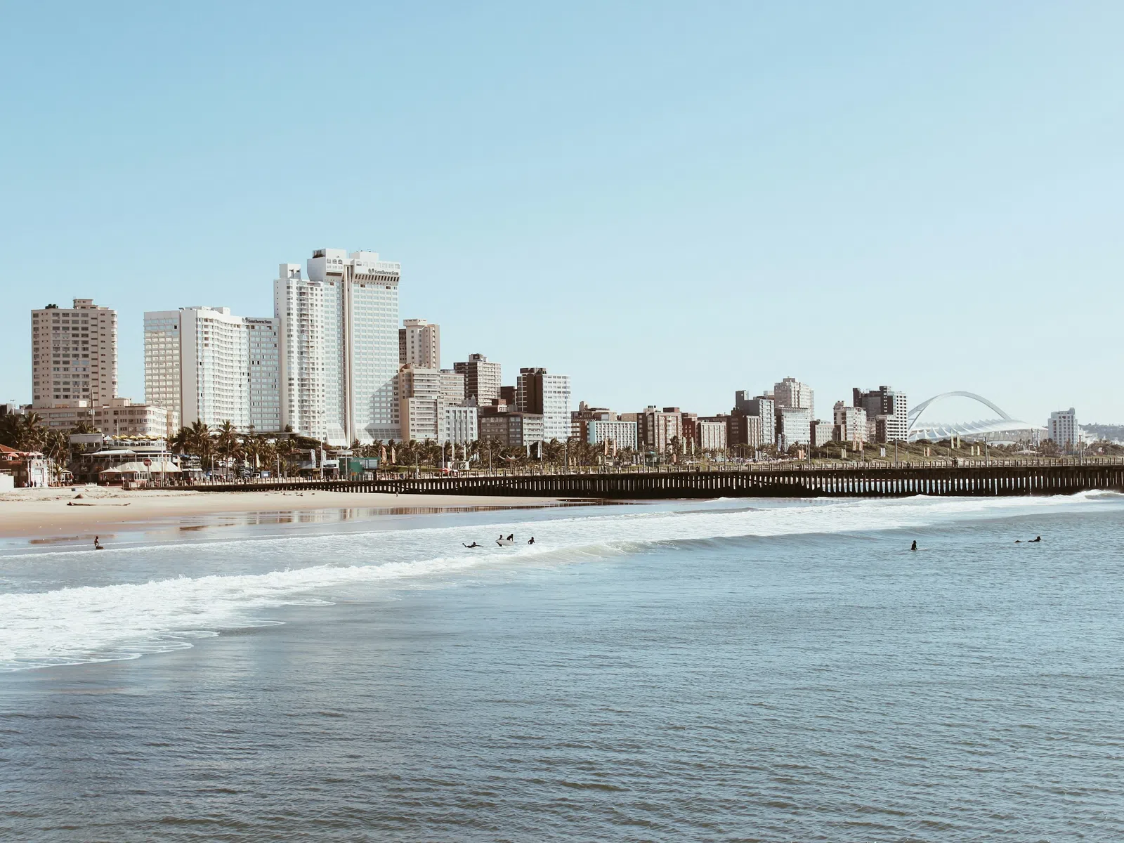 City skyline with tall buildings and beach shoreline with surfers and gentle waves under clear sky