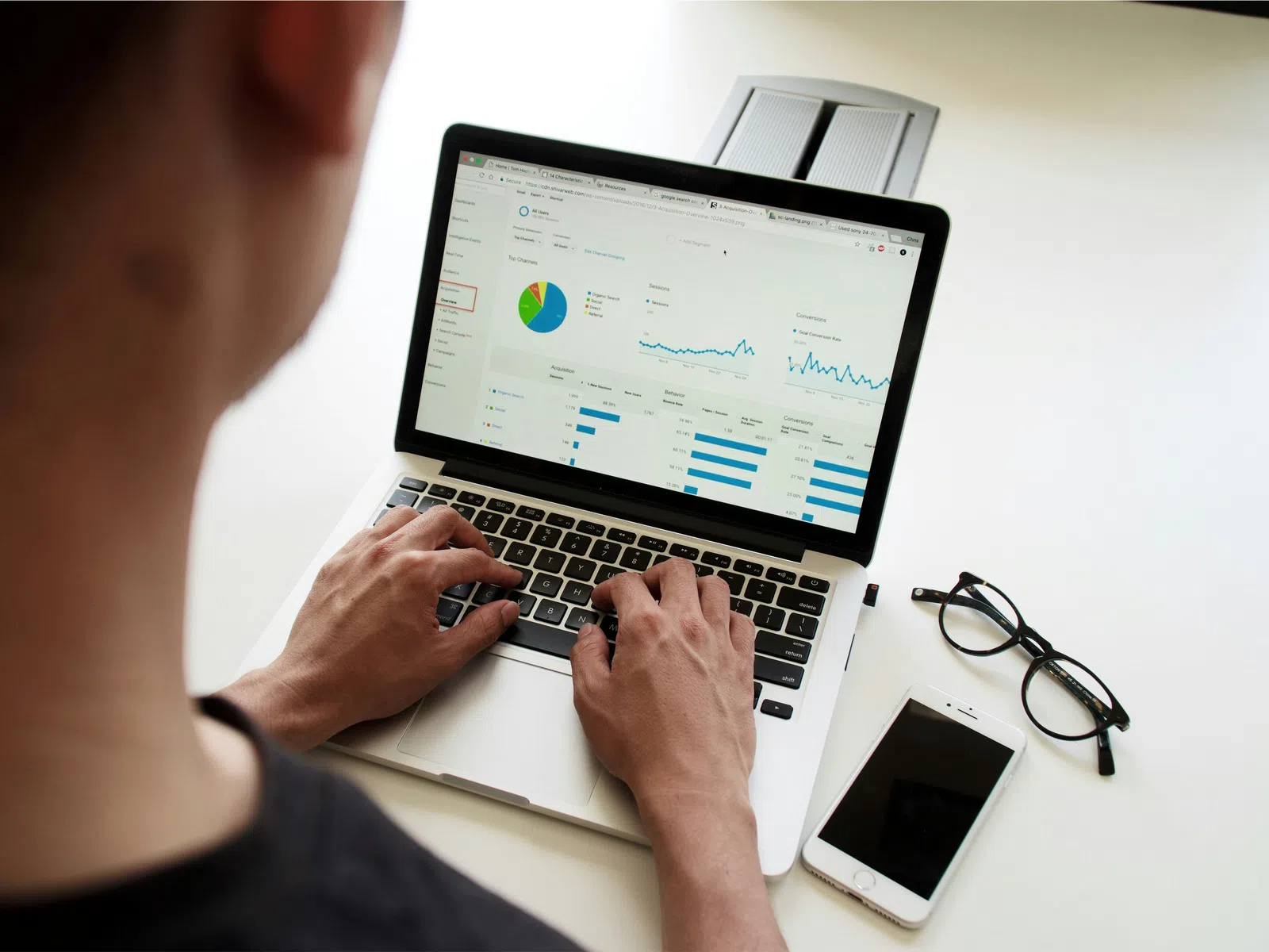 Person analyzing data charts and graphs on laptop with smartphone and glasses on white desk