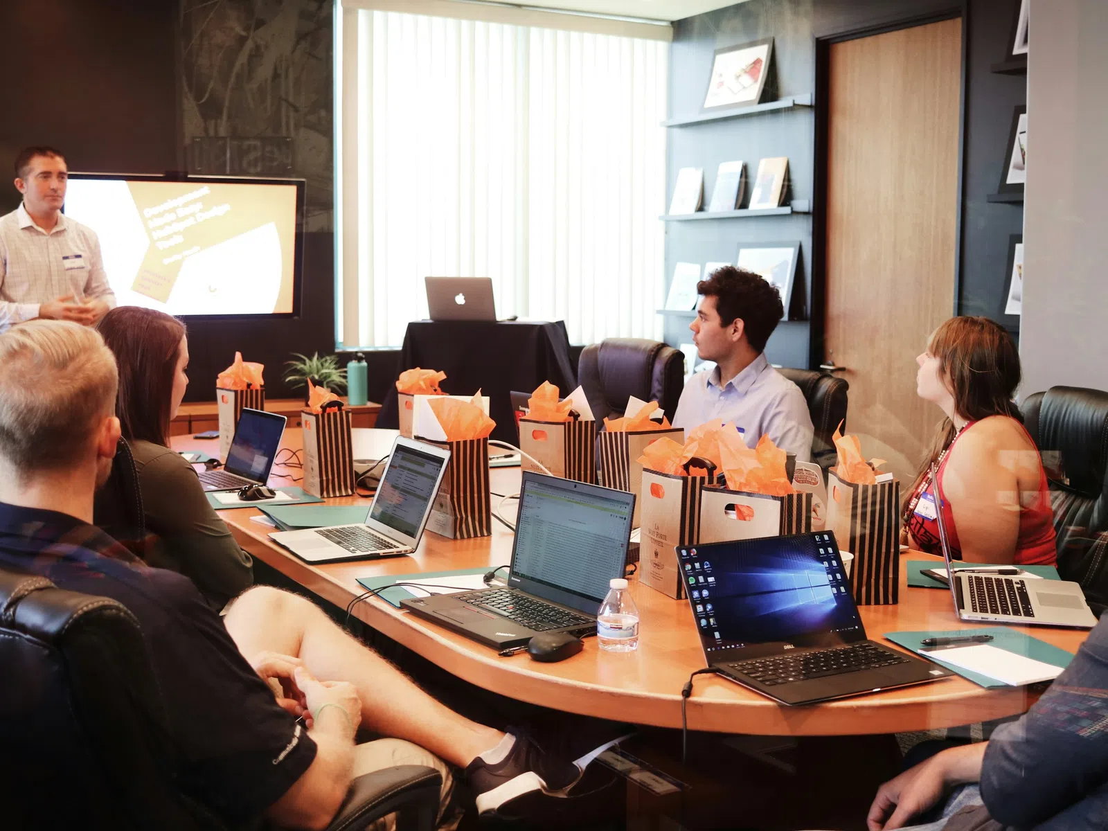 Business team in a meeting room listening to a presentation with laptops and gift bags on the table.