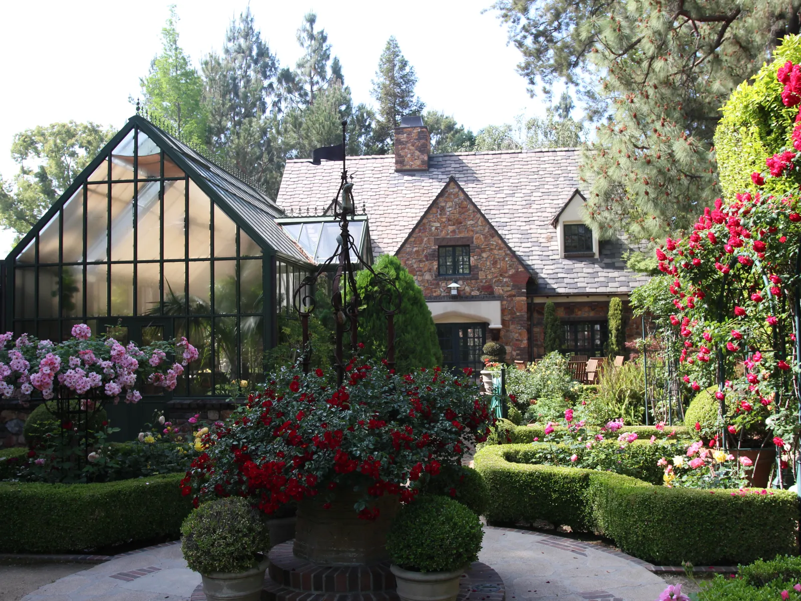 Charming stone cottage with glass greenhouse surrounded by vibrant rose bushes and manicured garden hedges.